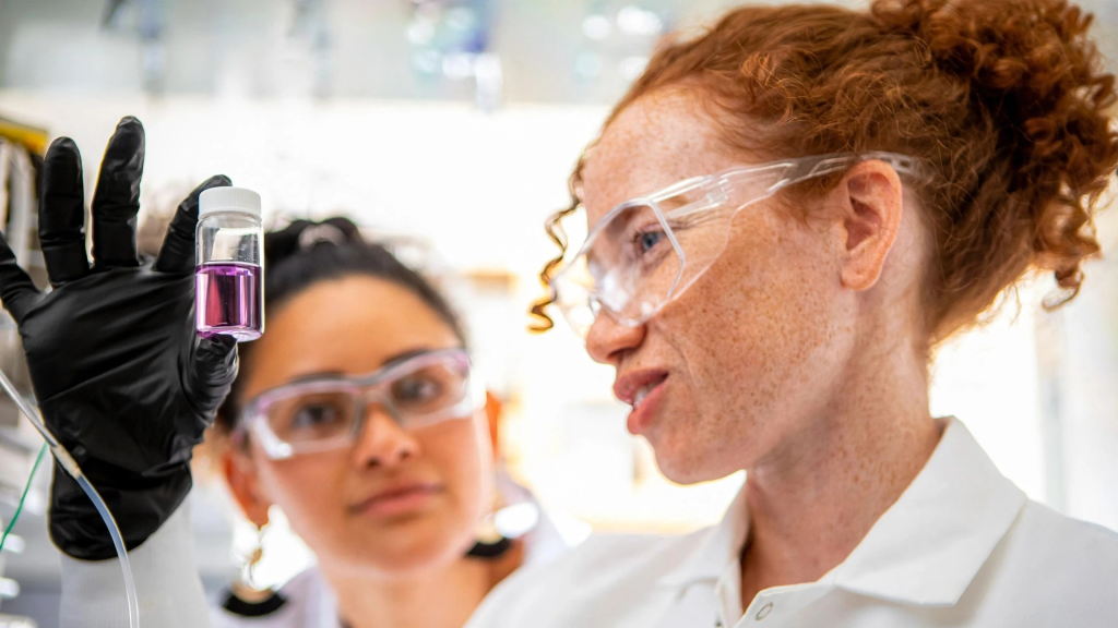 Twee laboratoriumanalisten met veiligheidsbril en witte labjassen onderzoeken aandachtig een monster in een glazen flesje met paarse vloeistof, representatief voor de expertise die nodig is bij de werving van laboratoriumpersoneel.